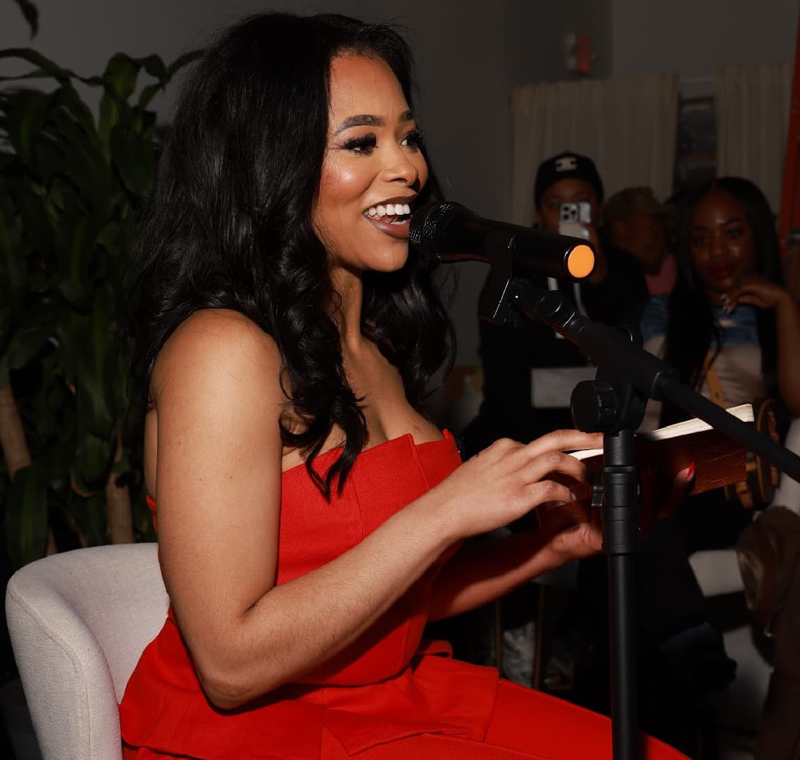 Alexus Brown reading poetry into a microphone at a live event, smiling, with an audience seated behind her