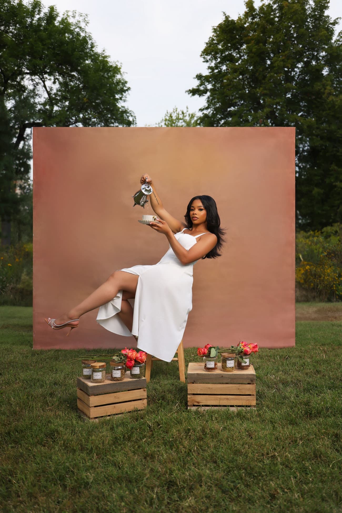 Alexus Brown, MoreLife Society's founder, seated and pouring tea outdoors, with labelled MoreLife tea jars on wooden crates beside her